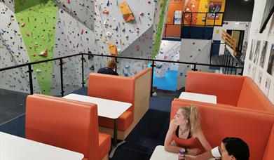 Two ladies sit on padded seats at a table in a café area on a balcony overlooking a huge part of the climbing walls.