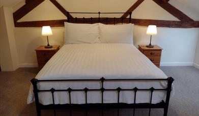 A black metal bedstead with comfy mattress and white bedding. There is a pine bedside table and table lamp to each side. Ceiling is sloping with expos