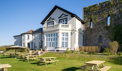 A Stunning White Hotel (The Castle Of Brecon Hotel) & The Medieval Castle Wall. The Sky Is Blue And there Are Light Wooden Benches On The Green Grass,