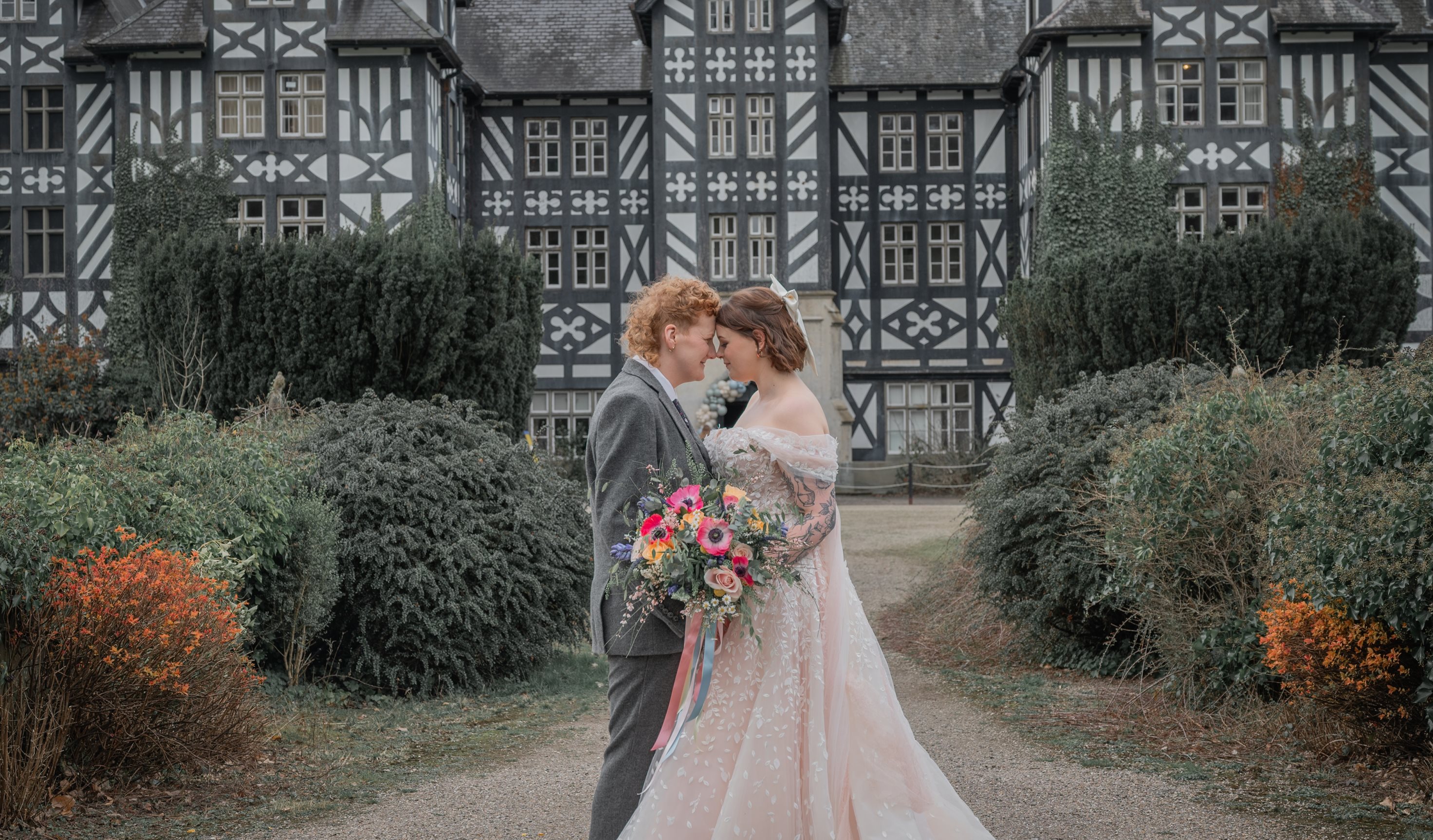 Two women in wedding outfits in front of a black and white stately home