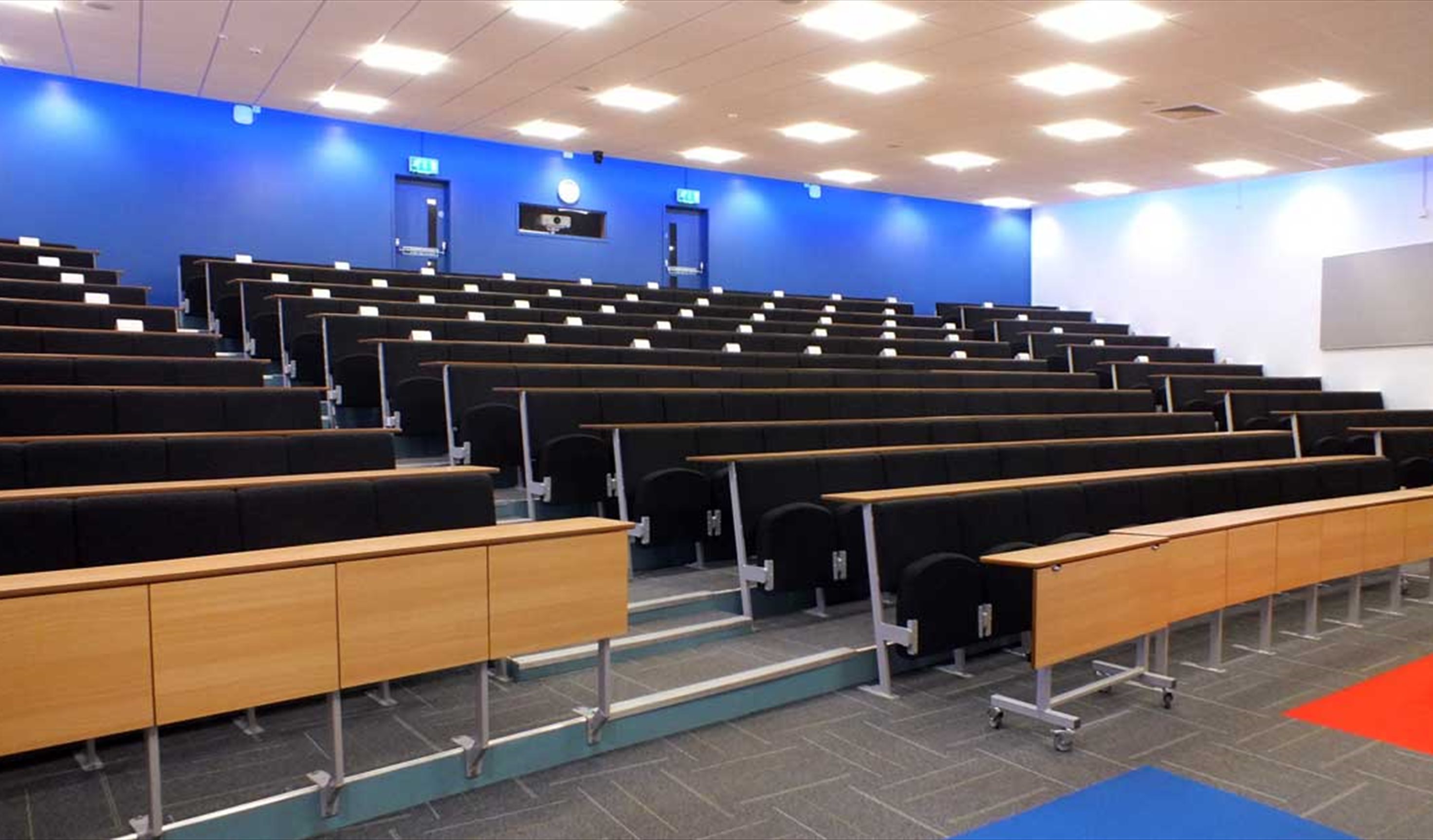 Empty lecture room with rows of seats and desks facing a screen.