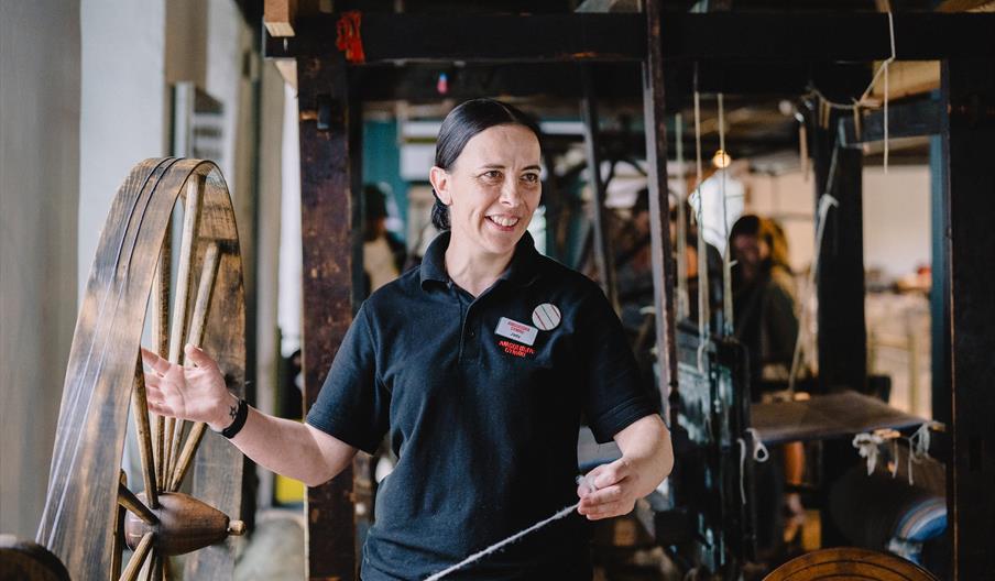 A woman dressed in a black polo shirt is spinning yarn on the Great Wheel in a gallery at the National Wool Museum