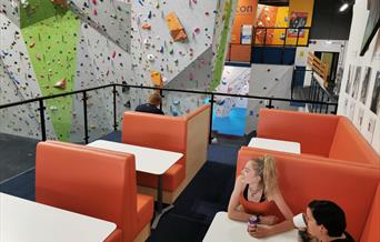 Two ladies sit on padded seats at a table in a café area on a balcony overlooking a huge part of the climbing walls.