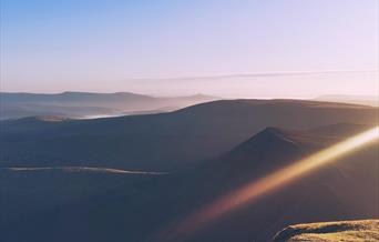 A scene of dawn overlooking layers of mountains of the Brecon Beacons, with golden highlights and pastel tones