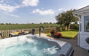 The beautiful, bubbling hot tub is in the foreground with the lawn stretching away. The patio dining table and chairs overlook the countryside