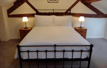 A black metal bedstead with comfy mattress and white bedding. There is a pine bedside table and table lamp to each side. Ceiling is sloping with expos