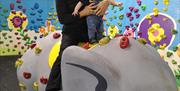 Grandad and toddler grandson sit on a manufactured elephant with climbing holds on it, and a little colourful climbing wall behind them in the play ar