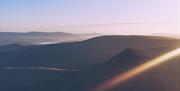 A scene of dawn overlooking layers of mountains of the Brecon Beacons, with golden highlights and pastel tones