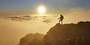 The silhouette of a walker on top of a mountain facing the sun rising above a cloud inversion.