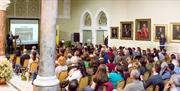 Rows of chairs with people watching a presentation. Grand setting with portraits and marble pillars