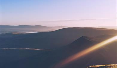 A scene of dawn overlooking layers of mountains of the Brecon Beacons, with golden highlights and pastel tones
