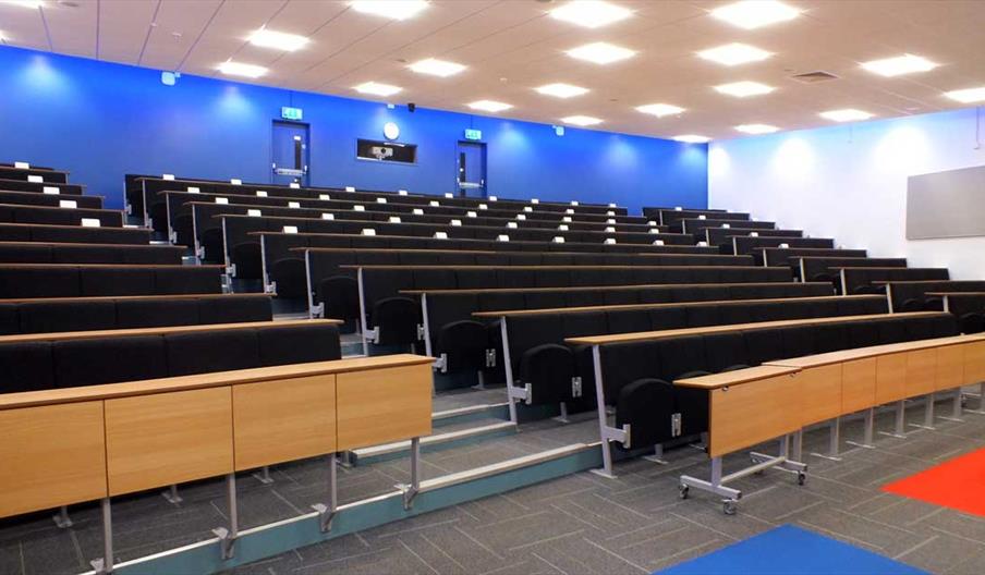 Empty lecture room with rows of seats and desks facing a screen.